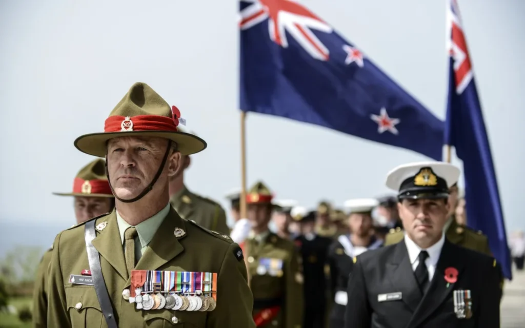 New Zealand military personnel wearing uniforms and medals during an Anzac Day memorial service, with the New Zealand flag waving in the background.
