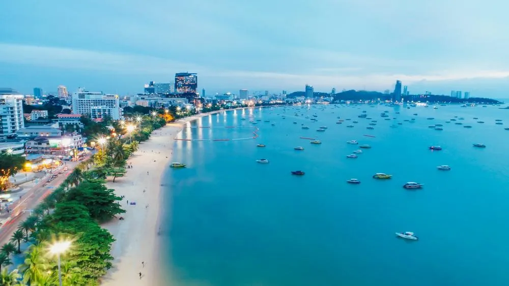 Beautiful aerial view of Pattaya Bay at twilight, showcasing the illuminated city skyline, a long curved sandy beach, and boats resting on the calm water.