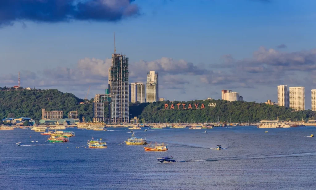 Scenic daytime view of Pattaya Bay showing numerous boats on the blue water, the iconic Pattaya City sign on the lush hill, and coastal skyscrapers.