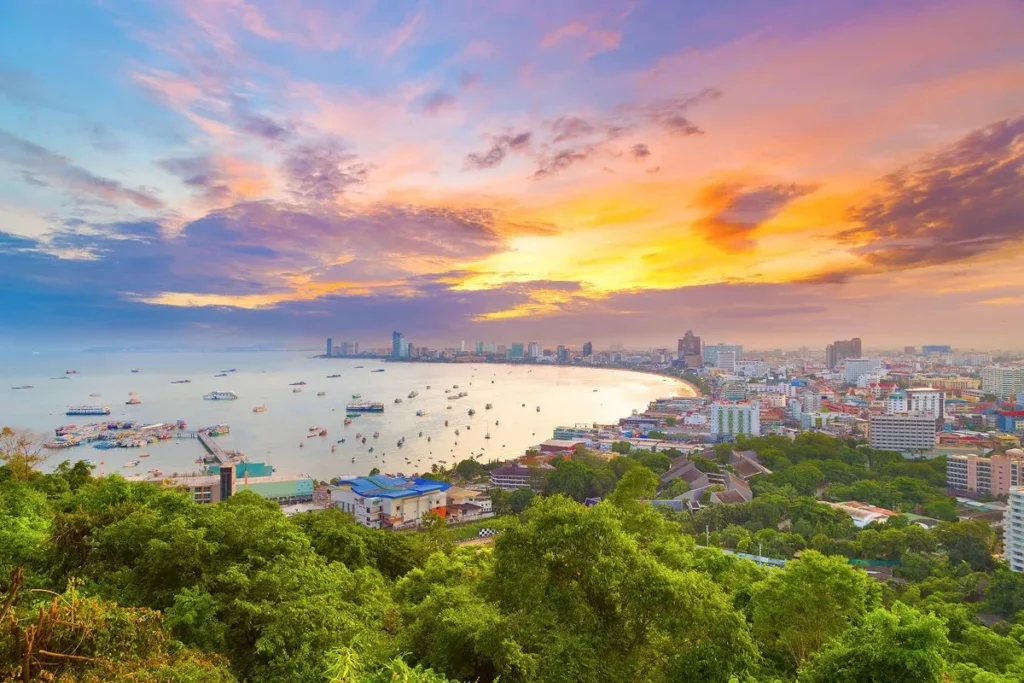 Panoramic elevated view of Pattaya Bay and the city skyline during a colorful sunset, showcasing the curved coastline, lush greenery, and boats in the ocean.