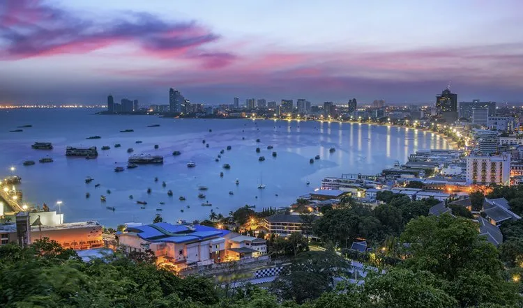 Panoramic viewpoint of Pattaya Bay during a stunning purple and pink sunset, highlighting the crescent-shaped coastline, glowing city lights, and boats scattered across the sea.