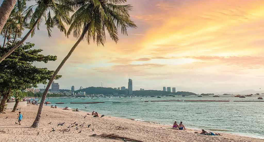 Scenic golden sunset over Pattaya Beach, featuring palm trees, pigeons on the sand, and people relaxing by the ocean.