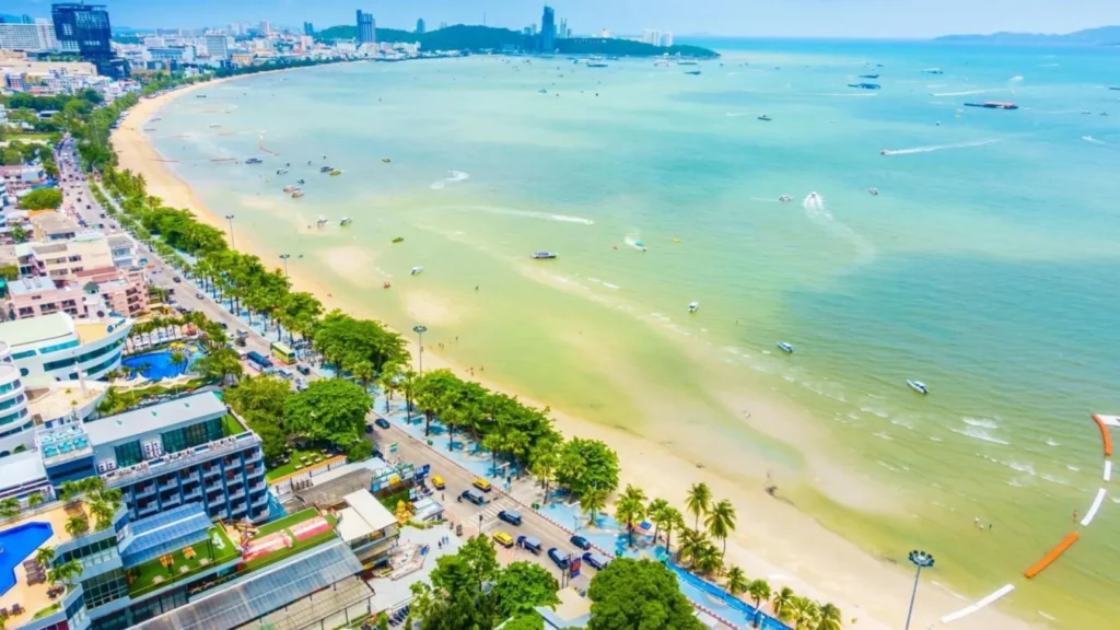 oramic shot of Pattaya beachfront road, green palm trees, sandy beach, and clear blue water with several speedboats.