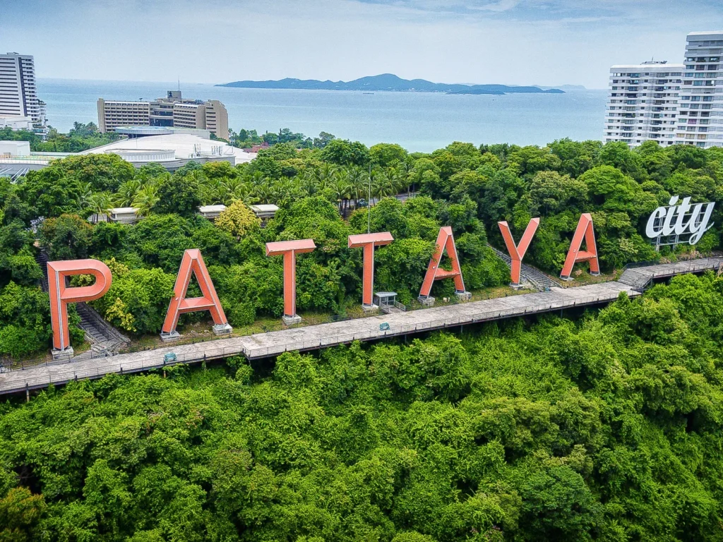 The iconic orange Pattaya City sign located on the lush green Pratumnak Hill, with a view of the ocean and coastal buildings in Thailand.