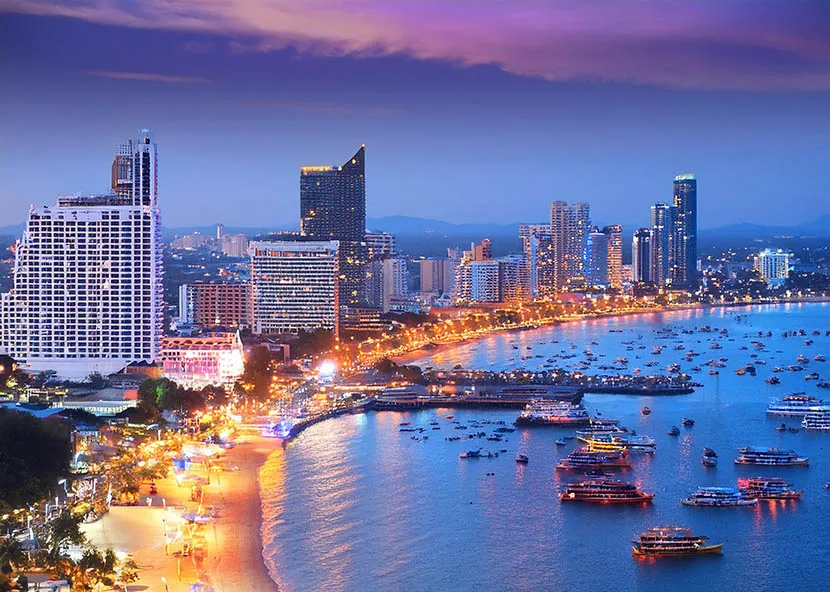 Vibrant night view of the Pattaya city skyline and beachfront, featuring brightly illuminated high-rise hotels and numerous boats anchored in the glowing bay.