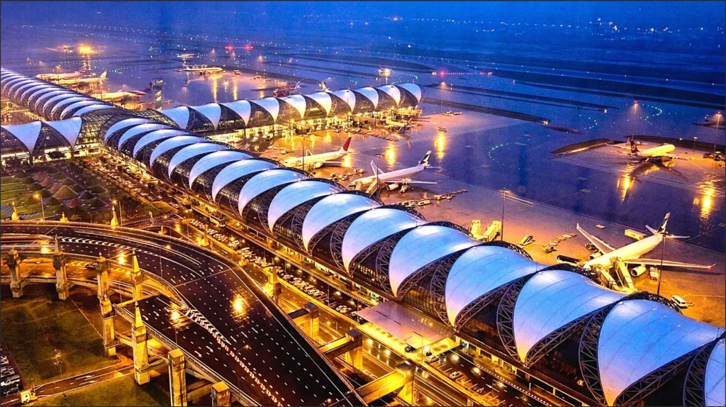 Aerial night view of Suvarnabhumi Airport in Bangkok, showcasing the brightly illuminated terminal building, concourse architecture, access roads, and airplanes parked on the tarmac.
