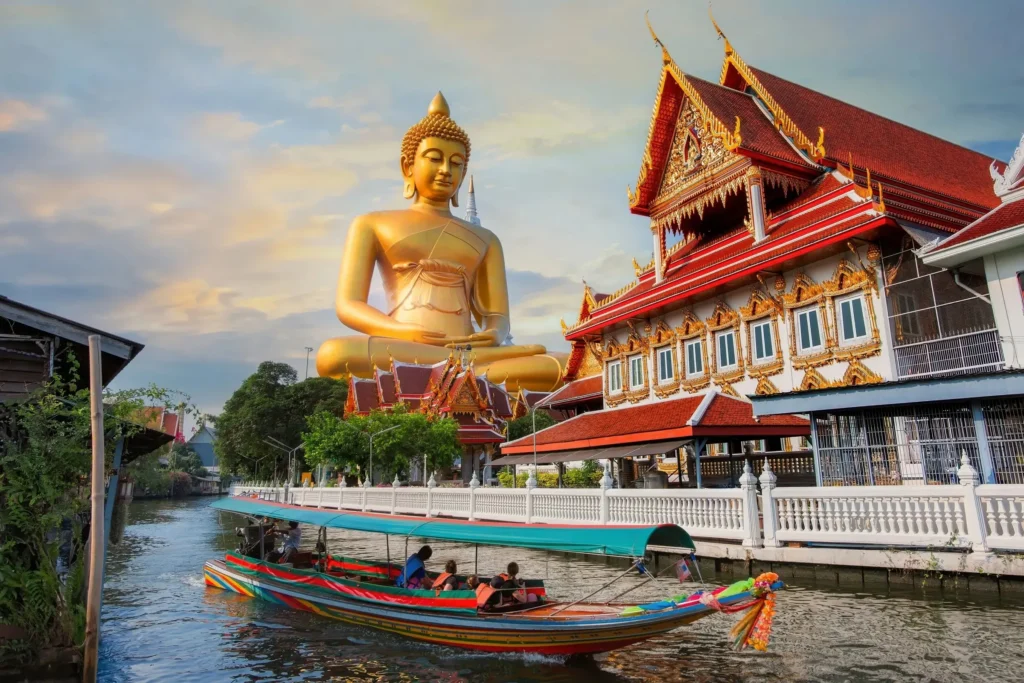Tourists riding a traditional Thai longtail boat on a canal with the spectacular Giant Golden Buddha of Wat Paknam Phasi Charoen in the background in Bangkok.