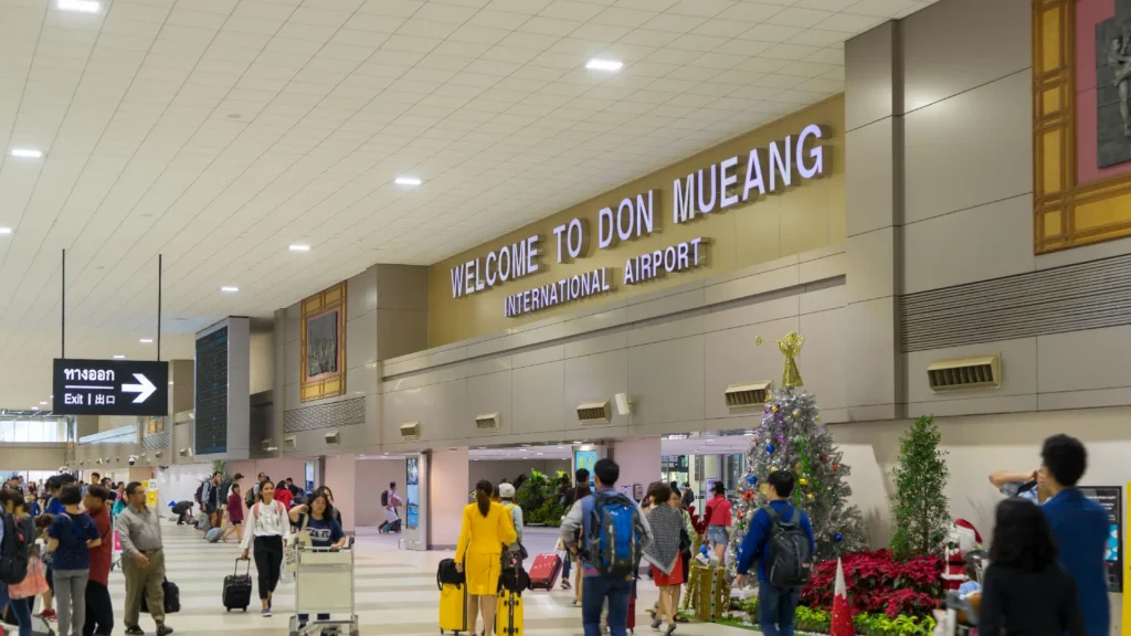 Passengers walking with luggage inside the terminal under the Welcome to Don Mueang International Airport sign in Bangkok, Thailand.