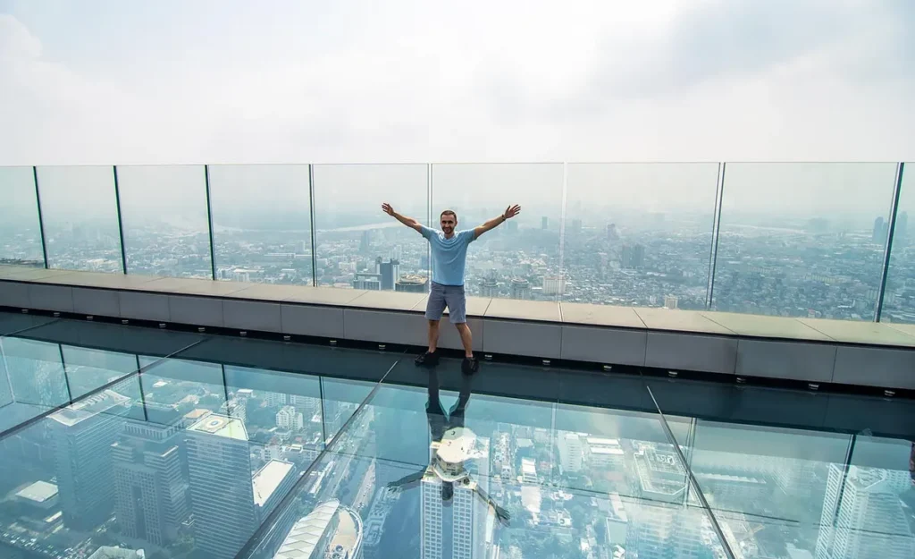Tourists sitting and taking photos on the thrilling transparent glass tray at the Mahanakhon Skywalk observation deck in Bangkok, Thailand.