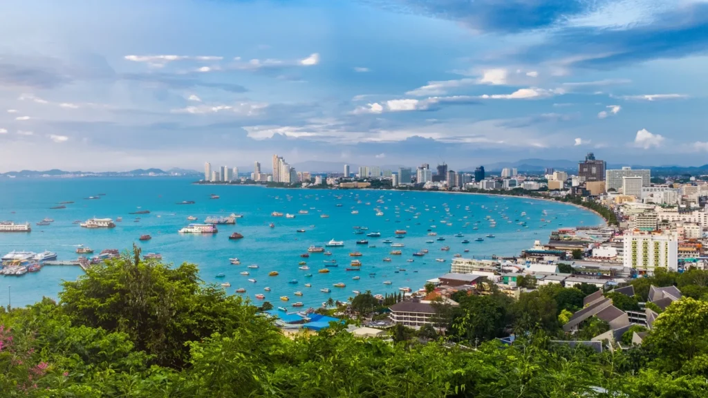 Stunning aerial panoramic view of Pattaya Bay, showcasing the curved coastline, beautiful blue ocean dotted with boats, and the modern city skyline under a partly cloudy sky.