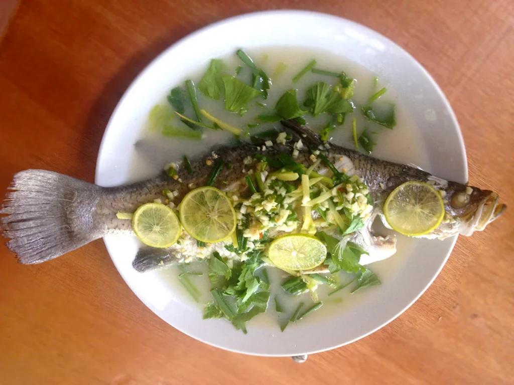 A white plate serving traditional Thai steamed fish topped with a flavorful and spicy clear broth made from fresh lime, garlic, and chopped red chilies.