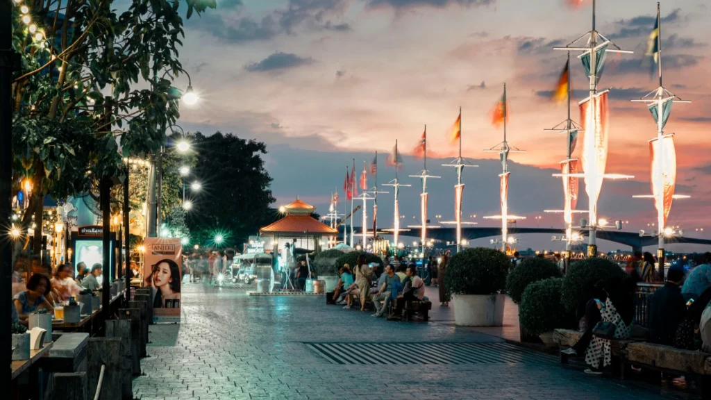 Picturesque sunset view over the Chao Phraya riverfront boardwalk at Asiatique in Bangkok, featuring decorative light poles and flags.