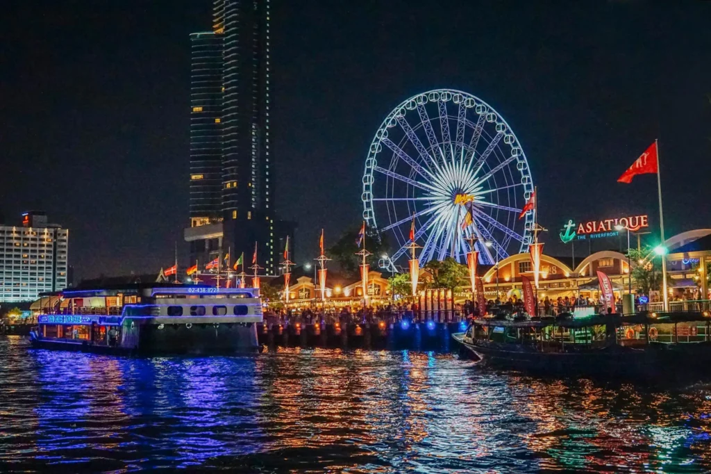 Night view of the illuminated Asiatique Ferris wheel and waterfront night market reflected on the Chao Phraya River with boats.