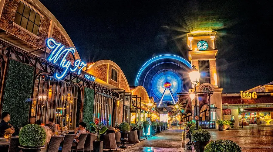 Vibrant night scene inside Asiatique The Riverfront showing the lit clock tower, outdoor restaurant seating, and a spinning Ferris wheel.