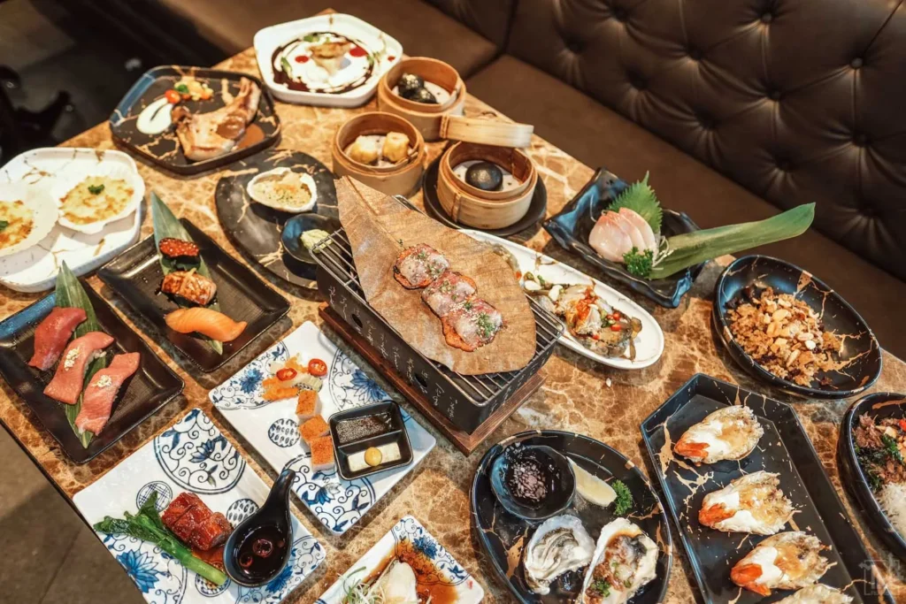 A lavish table spread of luxury Japanese and fusion cuisine featuring premium sushi, oysters, dim sum, and Wagyu beef on a tabletop grill.